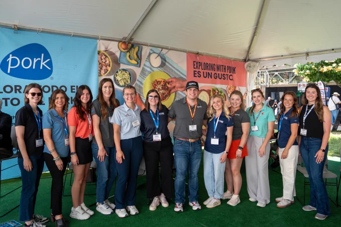A group of thirteen people stands under a tent, smiling at the camera. They are wearing casual attire with name tags and are posed in front of banners promoting pork, which feature images of various dishes and the phrase "Exploring with Pork es un Gusto." The atmosphere appears to be part of an event or exhibition.