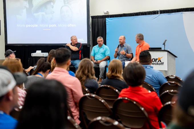 A panel of four men sits on a stage with a blue backdrop during a discussion at a conference. One man holds a microphone, engaging with a room full of attentive attendees. A large screen displays the words "More people. More pork. More often." Above the stage, a sign reads "Pork Checkoff."