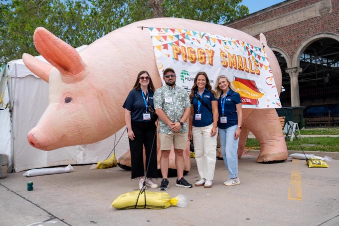 Four people stand smiling in front of a large inflatable pig with a banner that reads "Piggy Smalls" at an outdoor event. The background features a white tent and a brick building with an archway labeled "Fish and Game." The ground is secured with weighted bags, and trees and a blue sky are visible.