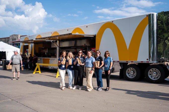 A group of six women stand smiling in front of a large food truck adorned with the iconic golden arches logo. They each hold brown paper bags and are wearing event badges. The truck is parked on a sunny street, with a clear blue sky and fluffy clouds above. A few people are visible near the truck, and a man carrying a bag walks nearby.
