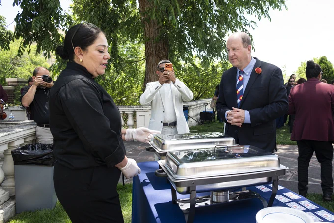 Caterer in black uniform serving food from chafing dishes at an outdoor event with guests mingling nearby under trees.