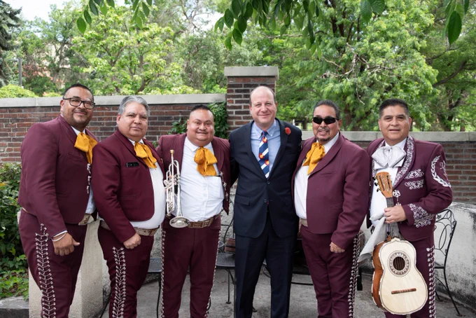Group of five mariachi musicians in traditional burgundy suits with yellow neckties posing outdoors with a man in a dark suit and striped tie.