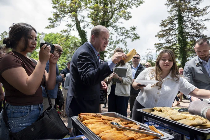 People gathered outdoors serving and receiving food from trays filled with empanadas under tall trees on a cloudy day.