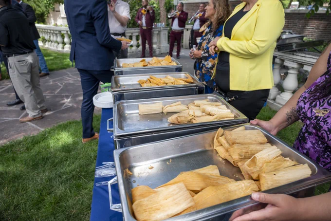 Chafing dishes filled with tamales lined up on a table at an outdoor gathering with people serving themselves.