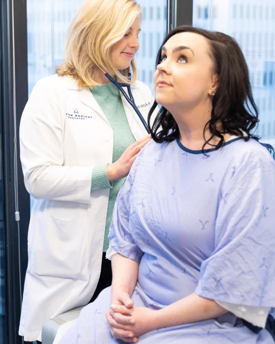 A person wearing a white coat and stethoscope examines a patient sitting on an exam table wearing a hospital gown