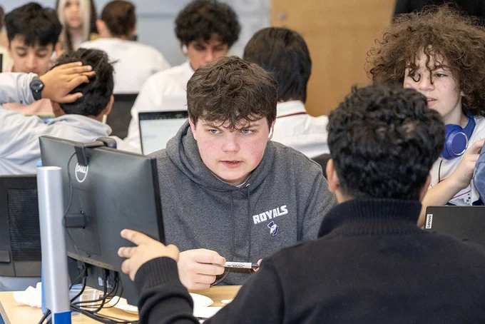 A group of students are seated in a classroom working on computers. One individual in the foreground is wearing a "Royals" sweatshirt and engaging in conversation with another person. There are several laptops and monitors on the desks.