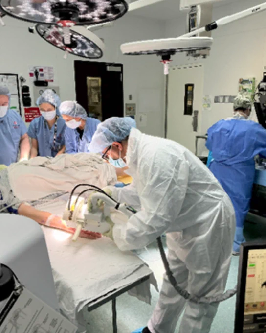 Medical professionals in a hospital operating room performing a procedure on a patient. They are all wearing surgical attire, including masks and gloves. The room is equipped with medical equipment and bright overhead lights.