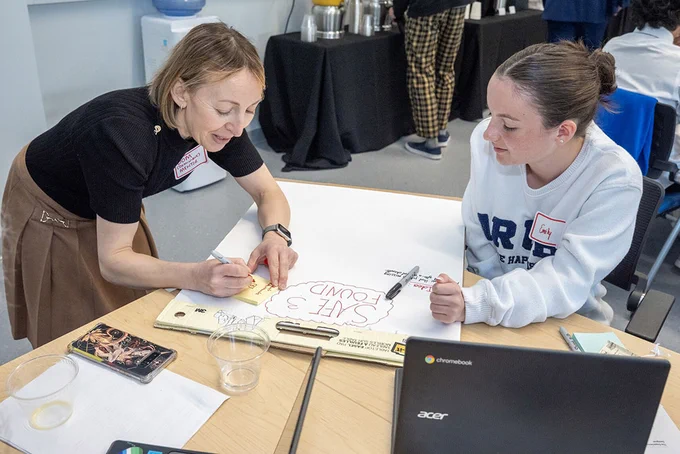 Two individuals are collaborating at a table. One is writing on a large sheet of paper with markers. An Acer laptop and several items, including a book and a plastic cup, are on the table. A water cooler and a refreshment table are visible in the background.