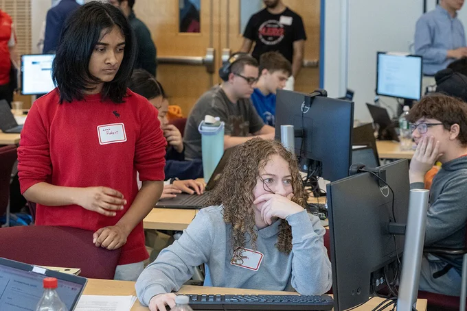 Participants in a computer lab work on screens, with others collaborating nearby.