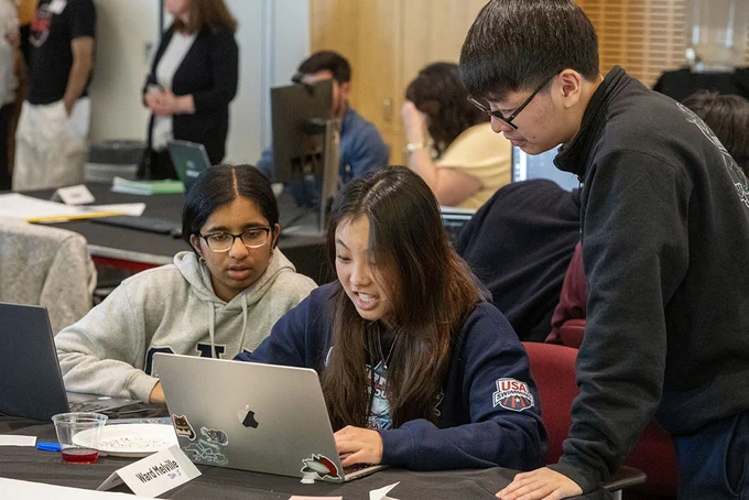 Three people are gathered around a laptop, engaging with the screen. One person is seated and typing, while the others look on with interest. There are various materials and a drink on the table, and other individuals can be seen in the background.