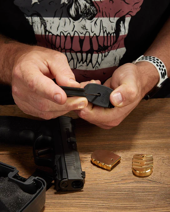 Hands installing a brass magazine base plate onto a SIG P365 X Macro magazine beside a pistol and additional brass mag plates.