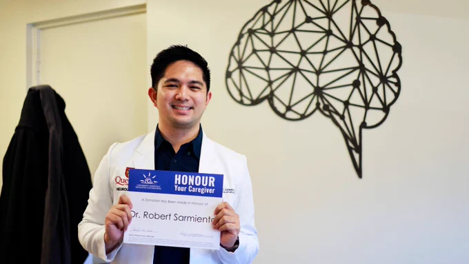 Dr. Robert Sarmiento smiling in a white Queen's jacket, holding an Honour Your Caregiver certificate with his name on it. Behind him on the wall is artwork of a brain.