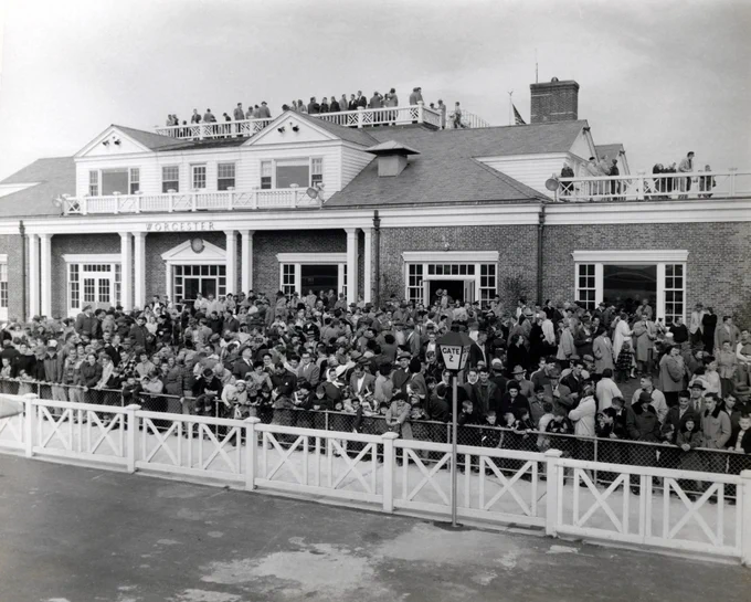 A historic image of a large crowd gathers outside the entrance of a traditional building with columns and a sign that reads "GATE." People are visible on the rooftop as well. The atmosphere suggests a public event or gathering.
