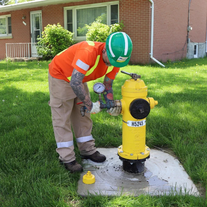 A person in safety gear, including a green hard hat and orange high-visibility shirt, is using a gauge on a yellow fire hydrant in a grassy area in front of a brick house.