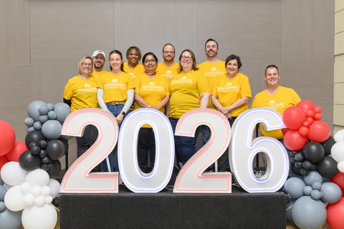 A group of people wearing matching yellow T-shirts stands behind a large display of the numbers "2026" surrounded by white, red, and gray balloons. The group smiles in a celebratory pose.