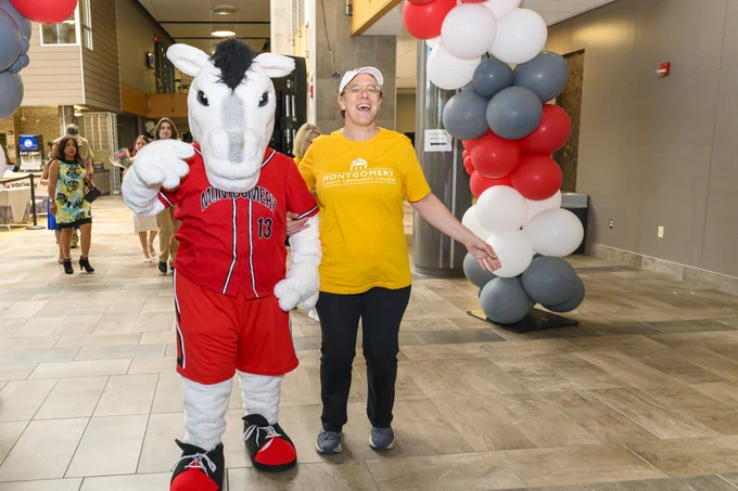 A person wearing a yellow shirt is standing alongside a horse mascot in a spacious hallway adorned with red, white, and gray balloon columns.