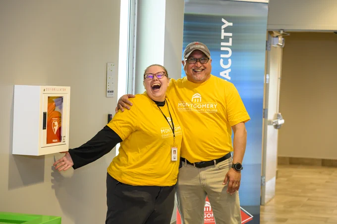 Two people smiling widely, each wearing a yellow "Montgomery County Community College" T-shirt. One has an arm around the other. They are standing indoors, near a wall-mounted defibrillator and signage with the word "FACULTY.