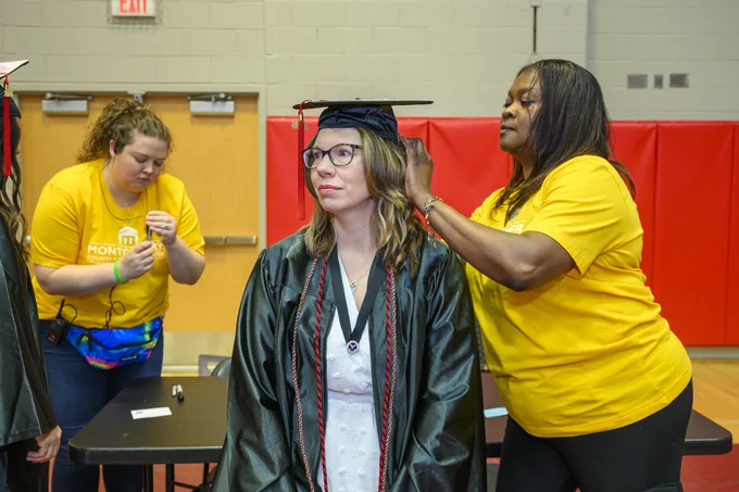 A person is wearing a graduation cap and gown, while another adjusts their tassel indoors. Both are in a room with red padding on the walls and a wooden door. Two others wearing yellow "Montco" shirts are seen in the background, one focused on a task.