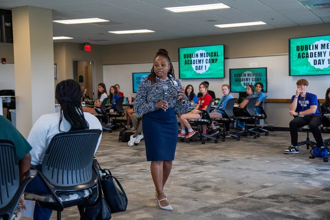 A presenter stands in the center of a classroom at the Dublin campus, speaking into a microphone to a room of high school students. Monitors in the background display "Dublin Medical Academy Camp Day 1."