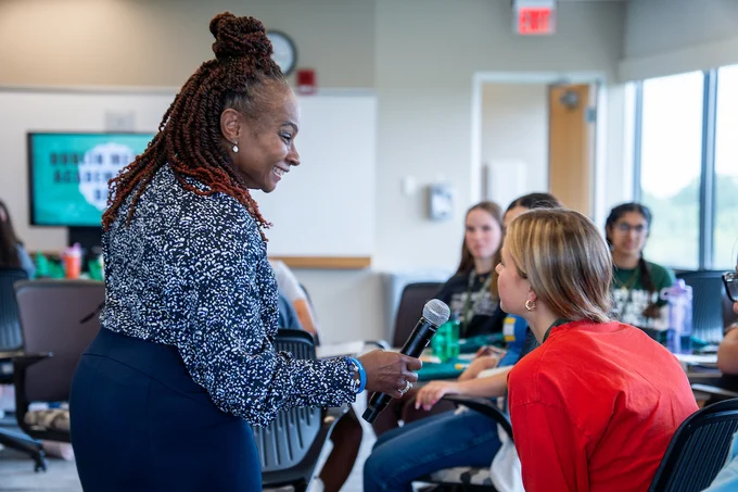 A presenter smiles as she holds a microphone for a high school student sitting at a desk during an interactive activity.