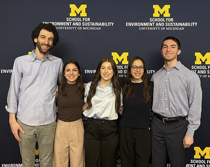 A group of five people standing in front of a backdrop with logos and text that read "School for Environment and Sustainability, University of Michigan." They are smiling and dressed in business casual attire.