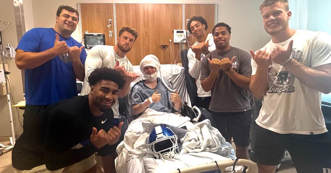 A group of people stand around a person in a hospital bed giving thumbs-up gestures. The person in the bed has a helmet on their lap. The setting looks like a hospital room.