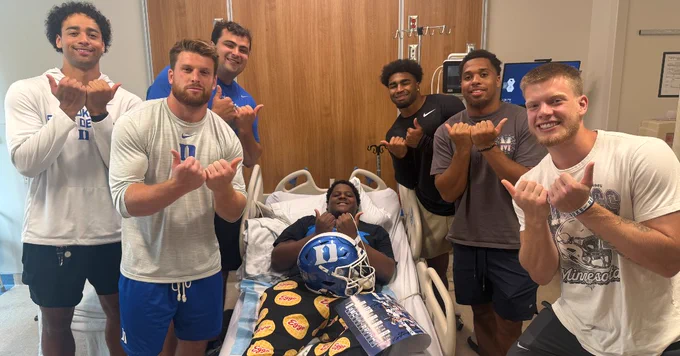 A person is in a hospital bed wearing a Duke football helmet, surrounded by six individuals making a gesture with their hands. The setting is a hospital room, and the Duke logo is visible on the helmet and a pillow.