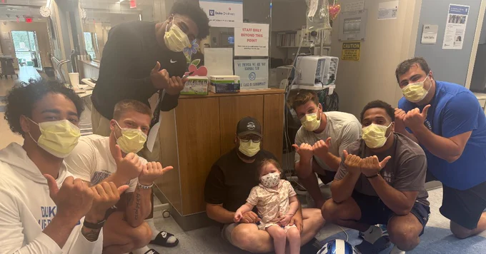 A group of people wearing masks pose together in a hospital setting. They are gathered around a small child sitting on the floor. Some of the individuals are wearing Duke Blue Devils apparel. There are medical carts and equipment visible in the background.