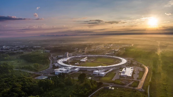 Aerial view of the Argonne National Laboratory during sunrise. The facility features a large, circular building surrounded by greenery and roads, with sunlight illuminating the horizon.