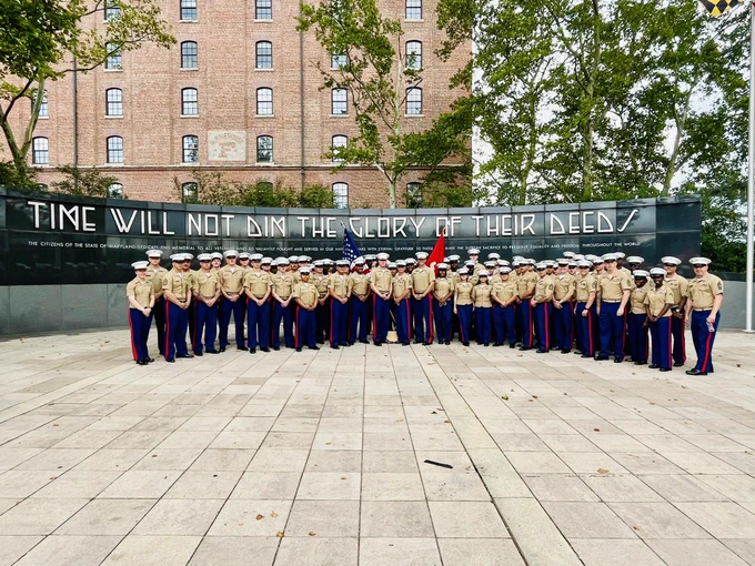Group of members of the military in uniform standing in formation in front of a wall with the inscription "TIME WILL NOT DIM THE GLORY OF THEIR DEED."