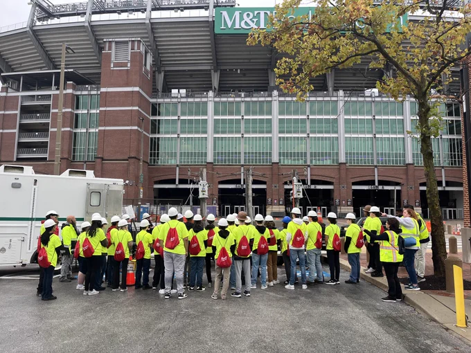 A group of students in construction hats in yellow and orange safety vests touring M&T Bank Stadium.