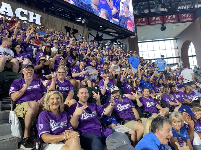 A large group of fans in purple "Rangers" jerseys sitting in a stadium, smiling and cheering. The background displays signs with years including 1996, 1998, 2010.