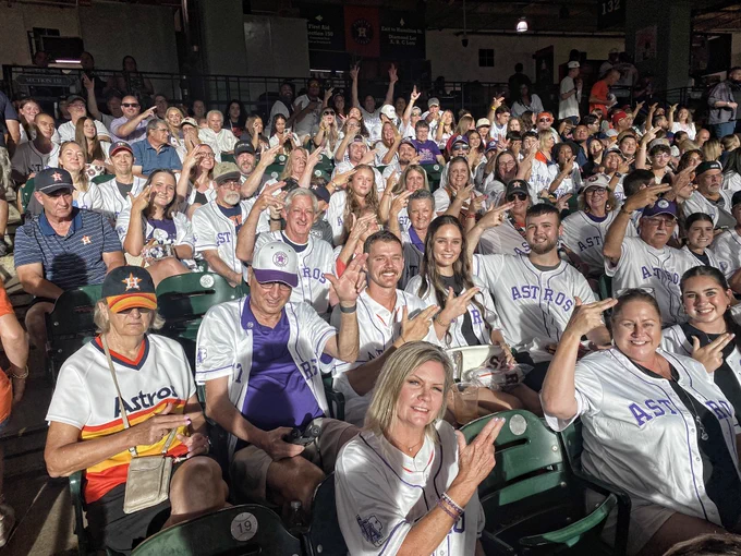 A large group of enthusiastic baseball fans wearing Astros jerseys pose for a photo in a stadium seating area. Many are smiling and gesturing with their hands, creating a lively and cheerful atmosphere.