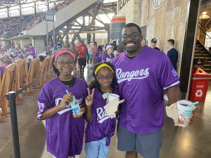 A group of three people wearing purple Texas Rangers jerseys is standing in a stadium hallway, smiling and holding cold treats. The area is bustling with other attendees in the background.