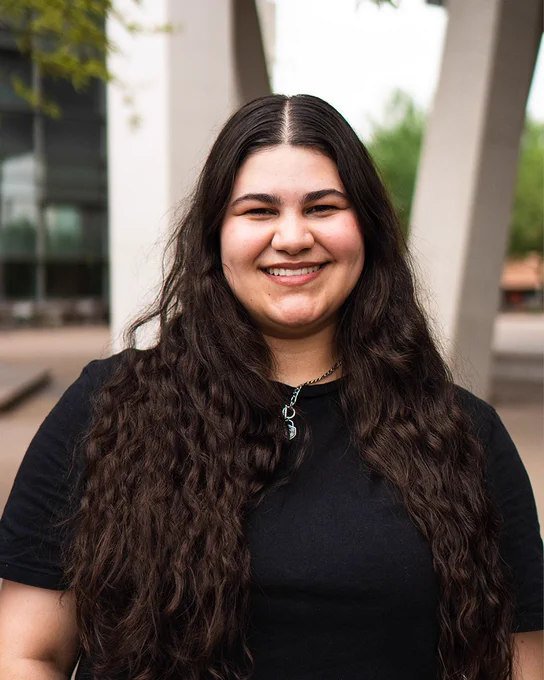 Alexandria Melick smiles at the camera while wearing a black shirt. Green foliage and building columns are visible in the background.