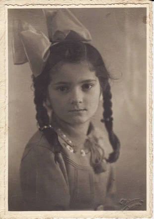 Vintage sepia-toned photograph of a young girl with braided hair and a large bow, wearing a necklace and a collared shirt, looking solemnly at the camera.
