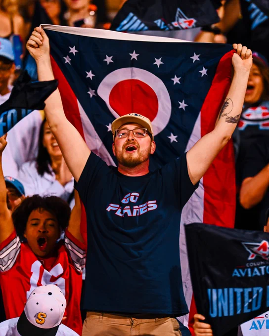 A group of enthusiastic fans cheer at a sporting event. One fan prominently holds up an Ohio state flag, while others wave team flags, creating an energetic atmosphere.