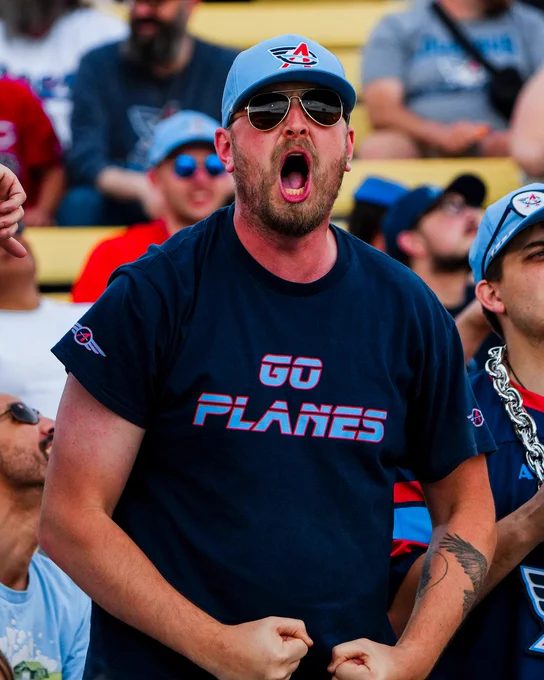 A passionate sports fan in the stands enthusiastically cheering. He is wearing a cap and sunglasses and has a tattoo on his arm.