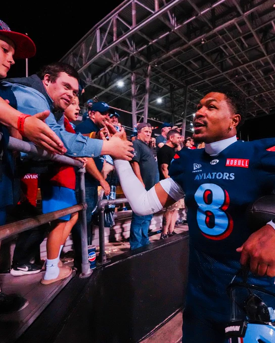A football player in uniform interacts with enthusiastic fans by the stadium stands. The scene conveys excitement and connection.