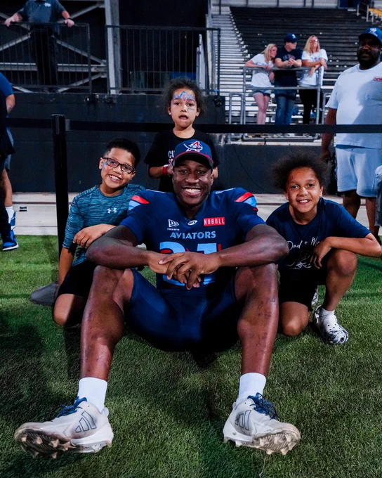 A football player in a blue and red uniform sits on the grass with three smiling children after a game. The atmosphere is joyful and celebratory.