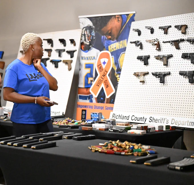 A person in a blue shirt stands looking at a display of firearms on a white pegboard. In the background, there's a banner with “Keenan” and a blue sports jersey. The table holds various firearm accessories. Text on the table reads “Richland County Sheriff’s Department.”
