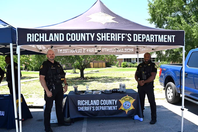 A booth set up by the Richland County Sheriff's Department. Two officers are standing in front of a tent displaying the department’s name. A table underneath holds various items, and a blue pickup truck is parked nearby. Trees and a building are visible in the background.