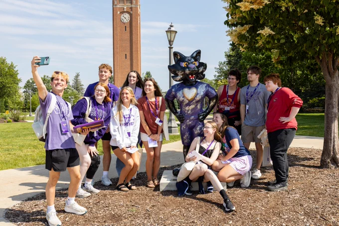 A group of individuals posing for a selfie with a TC statue on a college campus. In the background, there is a tall clock tower and a tree. The people are wearing lanyards and casual clothing, and the atmosphere appears to be lively and cheerful.