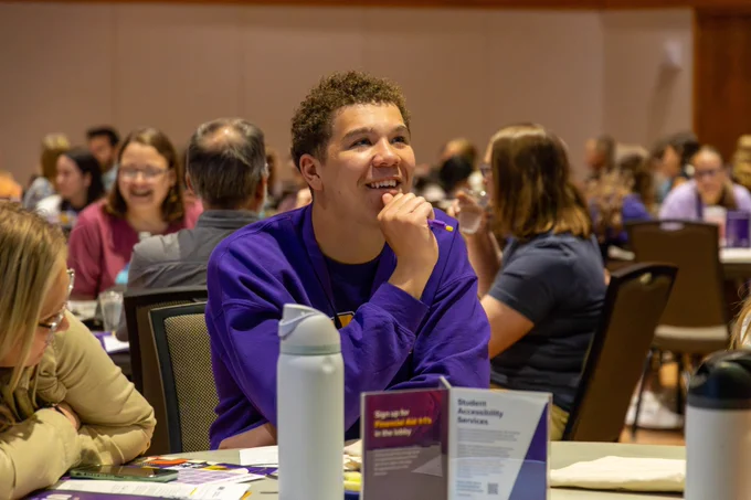 A person in a purple shirt sits at a table, smiling and holding a pencil. The table is covered with papers and a sign for Student Accessibility Services. The room is filled with people seated at various tables, engaged in discussion.