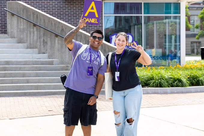 Two individuals smiling and standing on a campus walkway. One is holding a sign with the letter "A" and "DANIEL" printed on it. Both are wearing lanyards and casual clothing. There's a building and a 150 sign in the background. Flowering plants line the walkway.