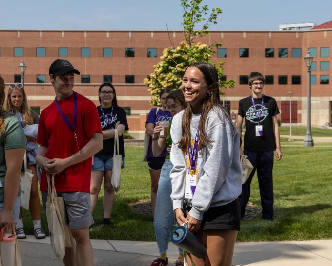 Several people are walking outside on a sunny day at a campus setting. One person in the foreground is wearing a gray sweatshirt. Others in the group are dressed casually and appear to be enjoying a tour. A brick building is visible in the background.