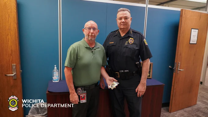 Two people stand in an office setting. One is dressed in a green polo shirt, holding an identification card, and the other in a Wichita Police Department uniform, holding a badge. The backdrop includes a blue partition, doors, plaques, and a hand sanitizer bottle. The Wichita Police Department logo is visible.