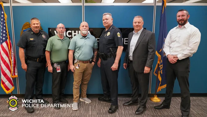 A group of six individuals in business and police attire stand in a room with blue partitions. An American flag is on the left, and there is a Wichita Police Department sign.
