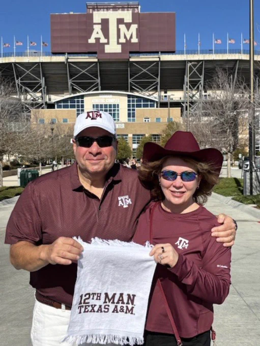 Two individuals are posing in front of the Texas A&M University stadium. They are wearing Texas A&M merchandise, and holding a "12th Man Texas A&M" towel.