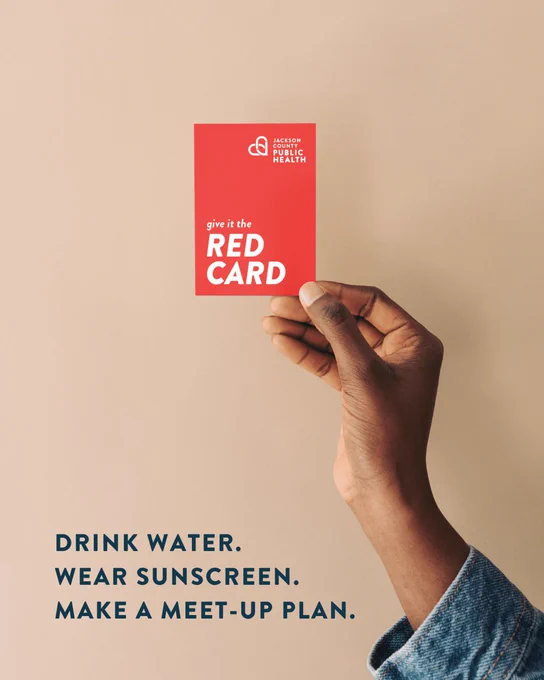 A hand holding a red card featuring the logo and name "Jackson County Public Health" with the words "give it the RED CARD." Below are the phrases: "Drink Water. Wear Sunscreen. Make a Meet-up Plan." on a plain background.